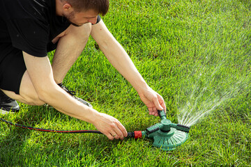 Male gardener adjusts a green lawn sprinkler on a vibrant green lawn, water spraying in arcs, showcasing a sunny outdoor setting with fresh grass © Anastasiia