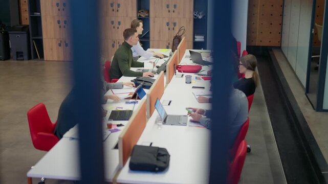 Diverse business professionals diligently work at their desks in a modern, collaborative office. Colleagues are focused on tasks, using laptops and writing, in a productive workspace.