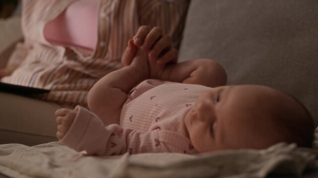 Rack focus shot of infant Caucasian girl wearing bodysuit lying on couch near unrecognizable mother working on laptop from home at night