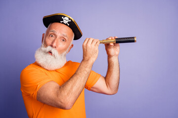 Older man in pirate hat peers through a telescope on a purple backdrop, wearing an orange shirt for...
