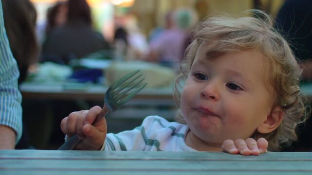 Toddler reaching across outdoor cafe table while holding fork and asking for food capturing curiosity, communication and authentic family mealtime behavior