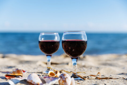 Two glasses of red wine on sandy beach with ocean horizon seashells and soft blur background summer relaxation travel lifestyle concept