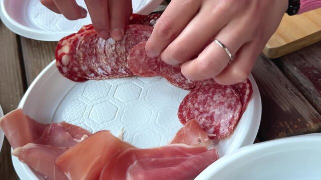 placing thin sausage slices and prosciutto onto white plastic plate during picnic table preparation with delicious meat serving atmosphere.