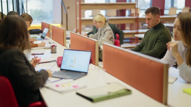 Business people are actively engaged in their tasks within a busy open-plan office. Employees concentrate on their laptops and tablets, collaborating and working efficiently in the shared workspace.