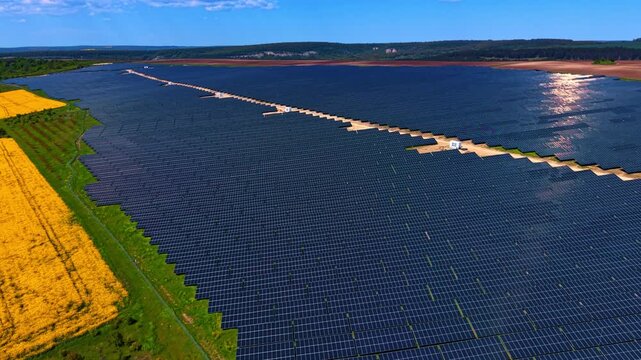 Footage over the huge field covered with modern shiny solar panels. Agriculture plantations surround the photovoltaic cell site. Aerial view.