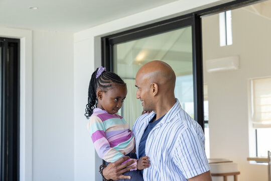 Father holding child daughter, smiling and looking down in bright living room near sliding door