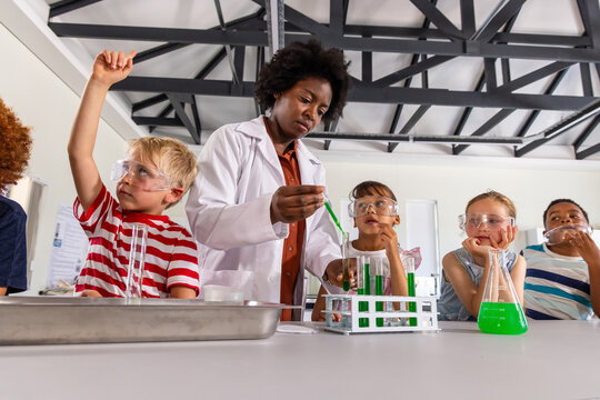 African American teacher in lab coat guiding children at lab table using dropper test tubes goggles