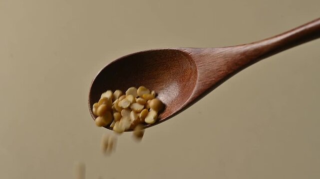 Dry yellow split dry peas gently falling from a wooden spoon against a neutral beige background. High quality close-up shot for culinary, cooking, and healthy nutrition concepts