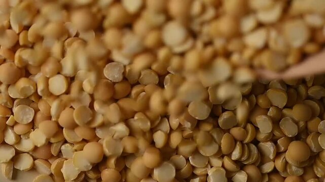 Close-up shot of dry organic split yellow peas pouring from a wooden bowl onto a heap against a beige background. Healthy vegan food ingredient. 
