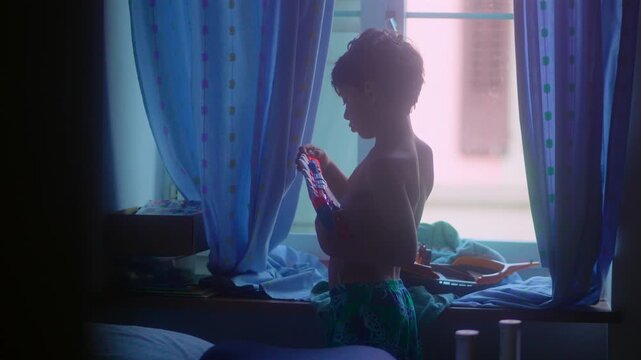 Young boy loading toy blaster in bedroom near window during playful indoor moment capturing imagination, childhood adventure and authentic everyday play at home