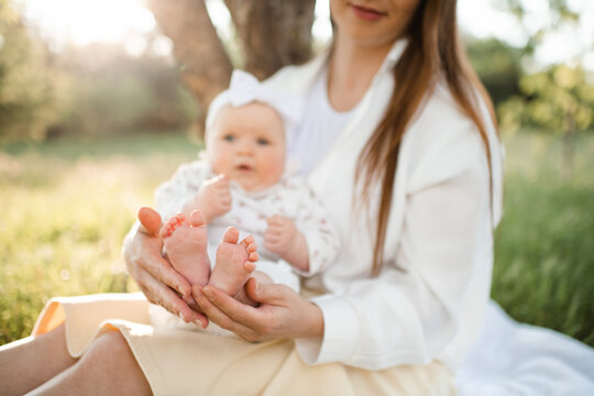 Happy woman holding baby feet sitting in green grass meadow under tree outdoors. Motherhood. Summer season.