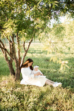 Happy woman holding baby sitting in green grass meadow under tree outdoors. Motherhood. Summer season.
