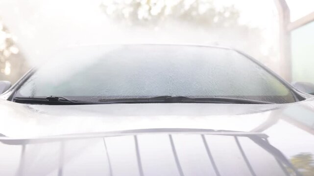 Water streams down a car's windshield during a cleaning process, with reflections visible on the hood
