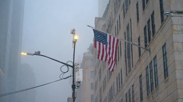 American flag on the building flaps in the wind under the falling snow. Terrible snow storm in New York City, USA. Low angle view.