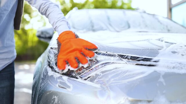 A person wearing an orange washing mitt cleans a car covered in soap suds, focusing on the headlight area