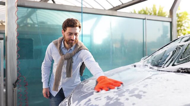 A smiling man wearing an orange glove washes his car with soap and water at a car wash station