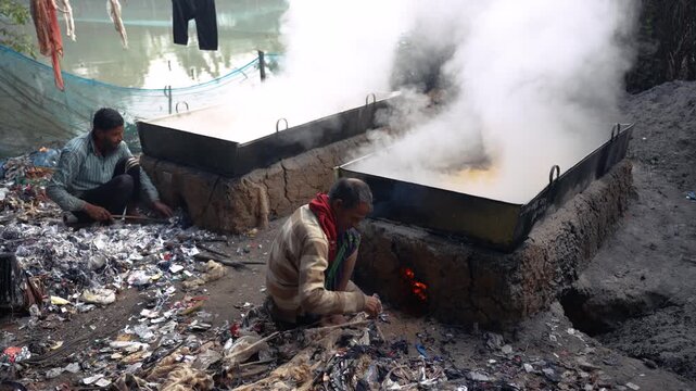 Natore, Bangladesh - 23 May 2026: View of workers tending to large metal vats boiling liquid over open mud stoves with thick white steam rising outdoors.