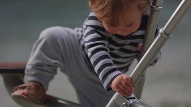 Toddler climbing onto playground digger structure while gripping metal handles capturing balance development, determination and authentic outdoor childhood exploration