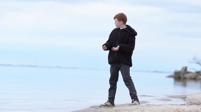 A teenager enjoys a fascinating game of throwing stones into the water of a lake, sea, or ocean. The boy relaxes, playing with stones and pebbles, throwing them into the lake. 