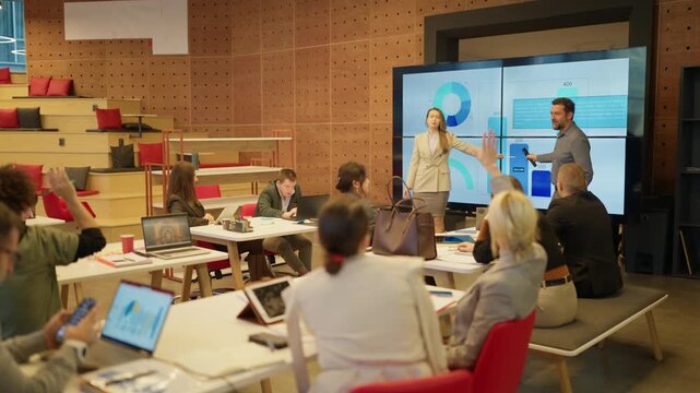 Businesspeople actively participate in a presentation featuring data visualization on large screens. Colleagues raise their hands during an engaging meeting in a contemporary workspace.