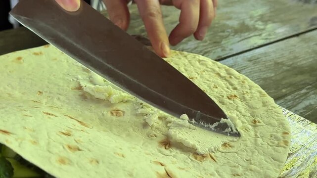 Homemade lavash snack preparation with creamy soft cheese being spread by knife across flatbread surface in detailed cooking atmosphere.