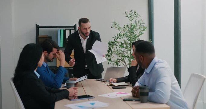 Portrait of furious Middle Eastern manager leans over table and points at coworkers during heated office meeting. Leader scolds team and tosses paper in anger