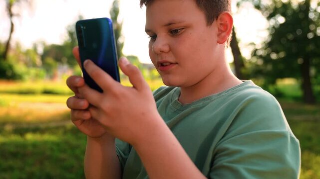natural background bokeh, youth using mobile internet outdoors, digital-native kid exploring apps outside in nature, boy concentrating on a phone screen near a meadow trail, green trees behind, boy