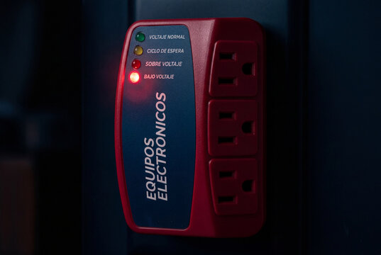 Primer plano macro de un protector de voltaje electr&oacute;nico enchufado, con la luz LED roja de alerta encendida en la opci&oacute;n de "Bajo Voltaje", sobre un fondo oscuro en penumbra.