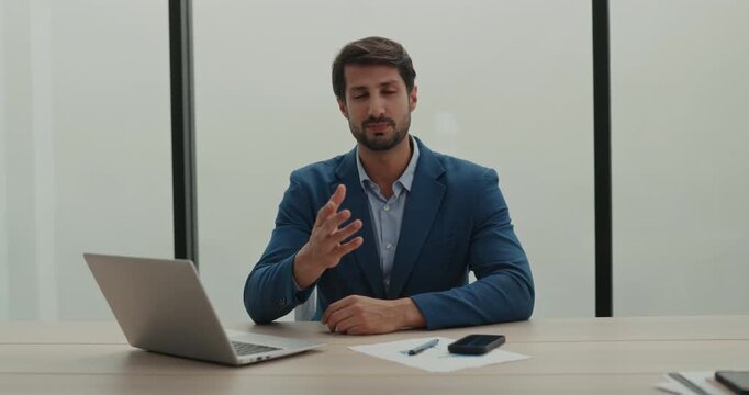 Portrait of Latin American finance manager explains strategy by laptop in office. Hispanic bearded business expert conducts online meeting in modern workspace