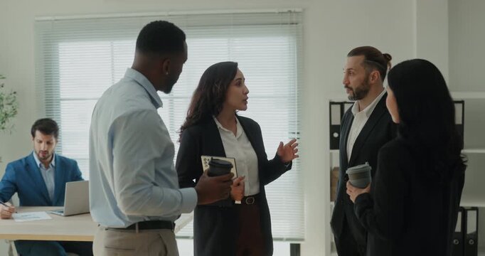 Portrait of positive Latin woman leader talks to multiethnic team in coworking. Young Hispanic female manager with tablet guides colleagues in modern office