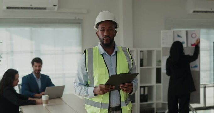 Portrait of African American construction engineer stands with tablet and looks at camera during work session. Specialist holds clipboard and waits for review