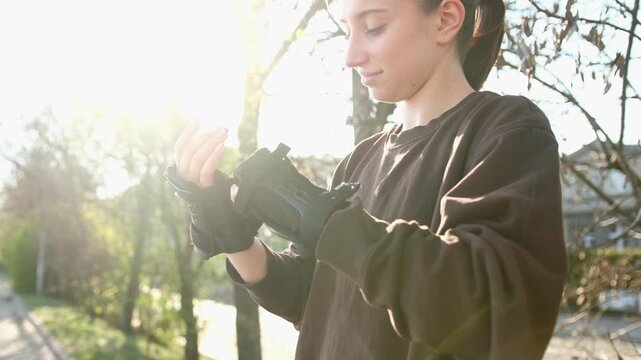 A girl puts on black wrist guards, preparing to roller skate. Sunlight shines through trees on a street sidewalk during the day.