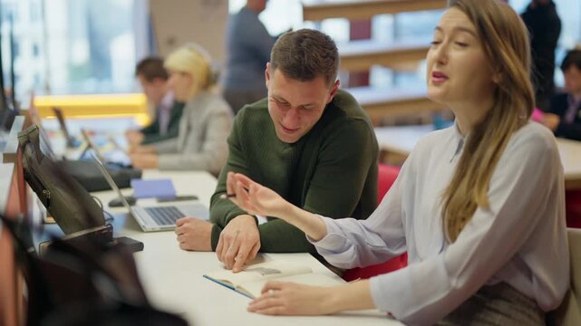 A young man and woman are discussing notes and sharing a laugh at a desk in a collaborative workspace. They are engaged in work, surrounded by other busy professionals.