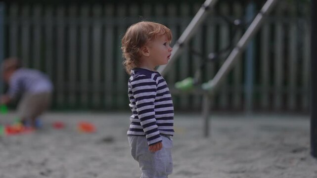 Toddler standing quietly in playground sandbox while observing surroundings capturing curiosity, awareness and authentic outdoor childhood exploration