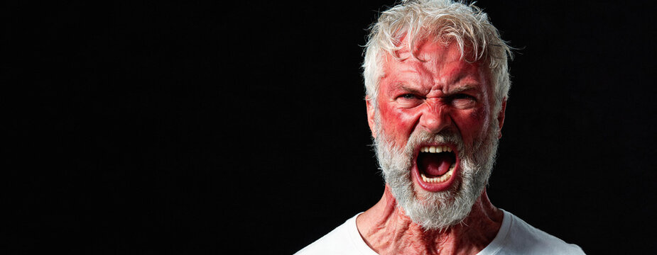 Senior man screaming with red face on black background. Man expressing aggression, frustration and extreme anger. Emotional portrait of furious senior man shouting loudly, wrath banner with copyspace.