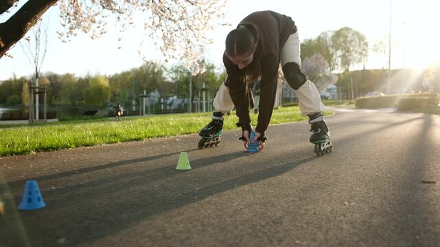 A young woman on inline skates places blue cones on a paved path in a sunny park. Golden evening light shines on trees and grass.
