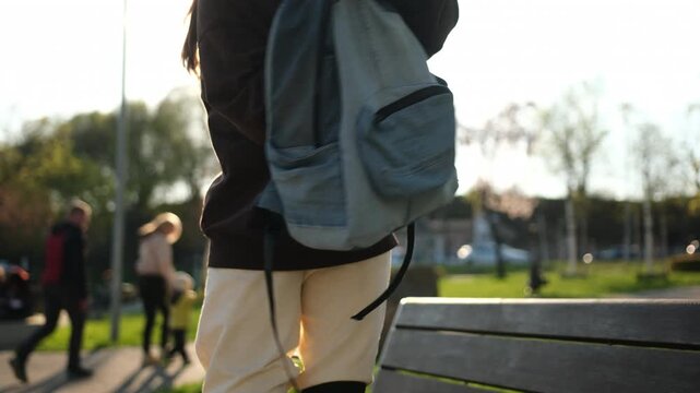 Girl on roller skates stands by a park bench, organizing her grey backpack. Sunlight shines as people walk in the park.