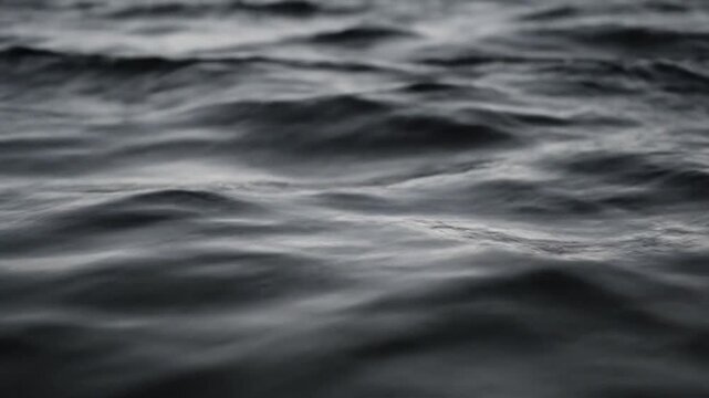 Dark and choppy deep blue water surface with swirling ripples and subtle light reflections. Close-up view of moody ocean waves moving in a rhythmic pattern during dusk.