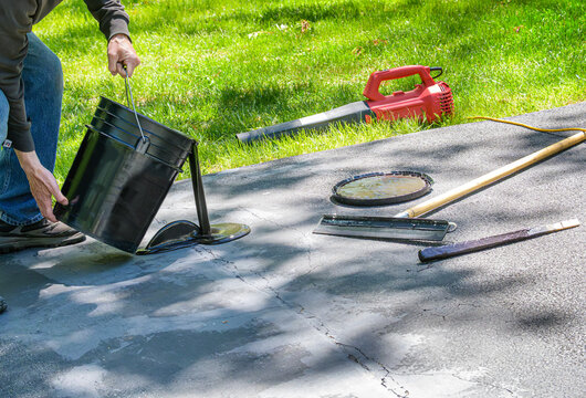 Man pouring blacktop sealer on driveway cracks with spreader and blower in background