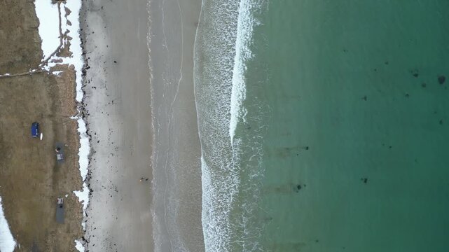 Aerial view of the turquoise waves crashing on the sandy beach with patches of snow Grotfjorden, Troms, Norway.
