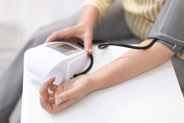 Fototapeta premium Senior woman measuring blood pressure at white table indoors, selective focus