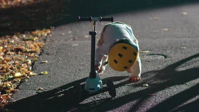 Toddler riding scooter after fall on sunny park path wearing safety helmet, young child learning balance and resilience outdoors, childhood development and confidence