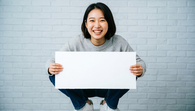 Smiling East Asian woman squatting and holding blank white banner against brick wall background