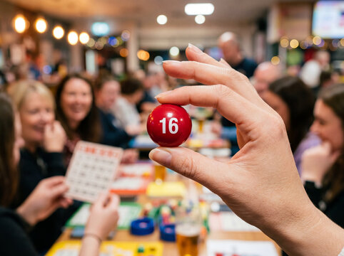 Hand holding a small red bingo ball displaying the number sixteen inside a busy community hall