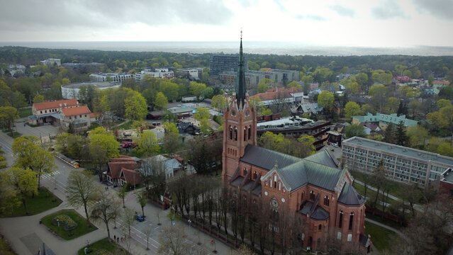 Aerial view of the Church of the Assumption of the Blessed Virgin Mary in Palanga, Lithuania. The Gothic Revival-style church features red brick construction and a tall, pointed spire. 
