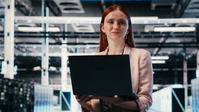 Portrait of smiling woman working in server farm doing network management virtualization, Cheerful data center female technician overseeing large scale IT infrastructure supercomputers with laptop.