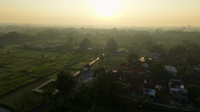 Aerial view of Plaosan Lor Temple surrounded by misty rice fields and village rooftops during a golden sunrise in Kecamatan Prambanan, Jawa Tengah, Indonesia.