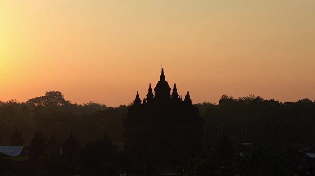 Aerial view of the silhouette of Plaosan Lor Temple against a golden sunrise sky with misty trees in Kecamatan Prambanan, Jawa Tengah, Indonesia.
