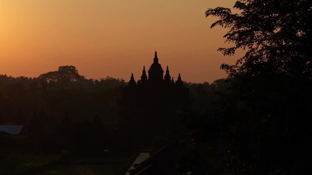 Aerial view of the silhouette of Plaosan Lor Temple against a vibrant orange sunset sky with trees and mist in Kecamatan Prambanan, Jawa Tengah, Indonesia.