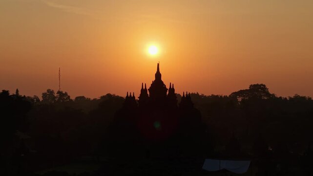 Aerial view of Plaosan Lor Temple silhouetted against a vibrant orange sunset sky in Kecamatan Prambanan, Jawa Tengah, Indonesia.